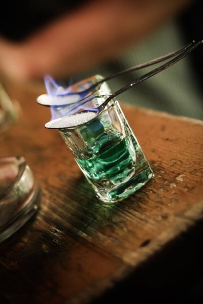 a pair of tongs holding a shot glass with a blue liquid and a flame