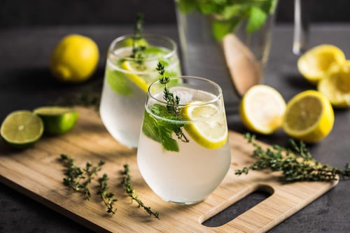 two glasses of liquid with lemons and herbs on a cutting board