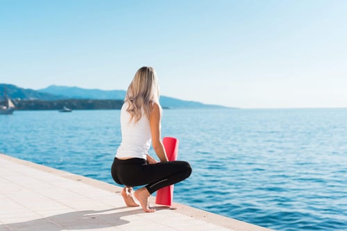 a woman kneeling on a ledge with a red yoga mat by the water