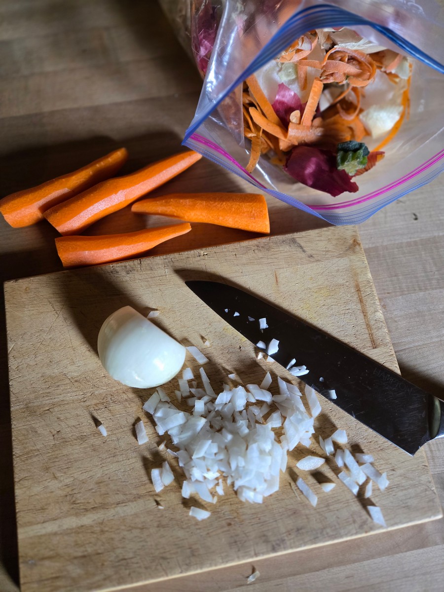 a bag of saved vegetable scraps and peels sits in a freezer bag next to a cutting board where onions are being chopped and carrots are waiting their turn.