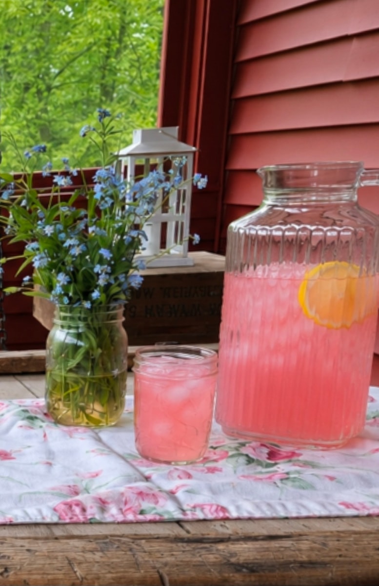 close up of rhubarb lemonade in glass pitcher sitting next to a jar of fresh cut forget me not flowers