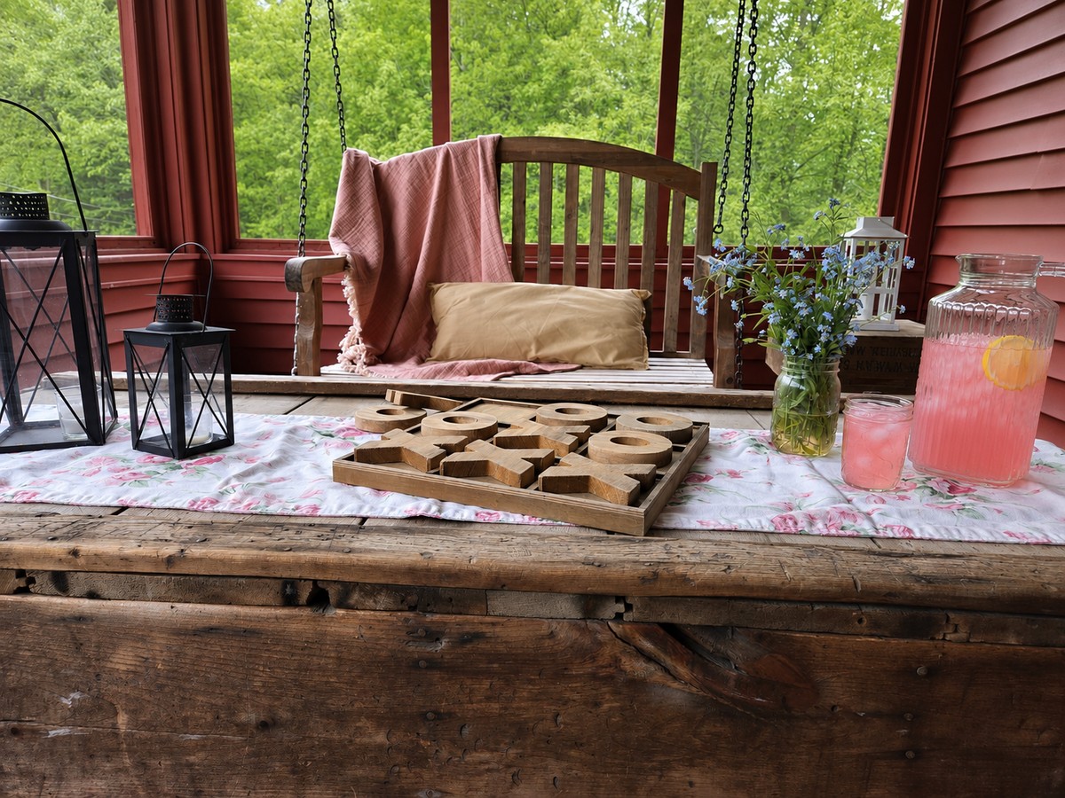 summer on the porch with a glass pitcher of rhubarb lemonade. A porch swing sits in the background, while the rhubarb lemonade sits on an old wooden trunk next to a wooden tic tac toe game board.