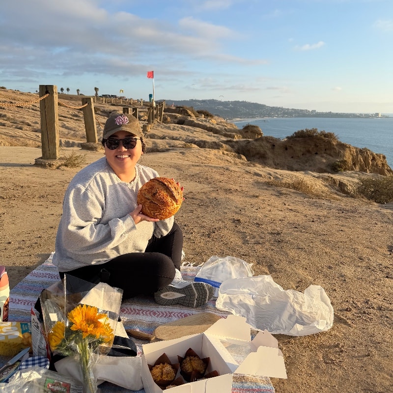Asian female home baker smiling and holding a loaf of sourdough bread, sitting next to sunflowers and muffins at the beach. 