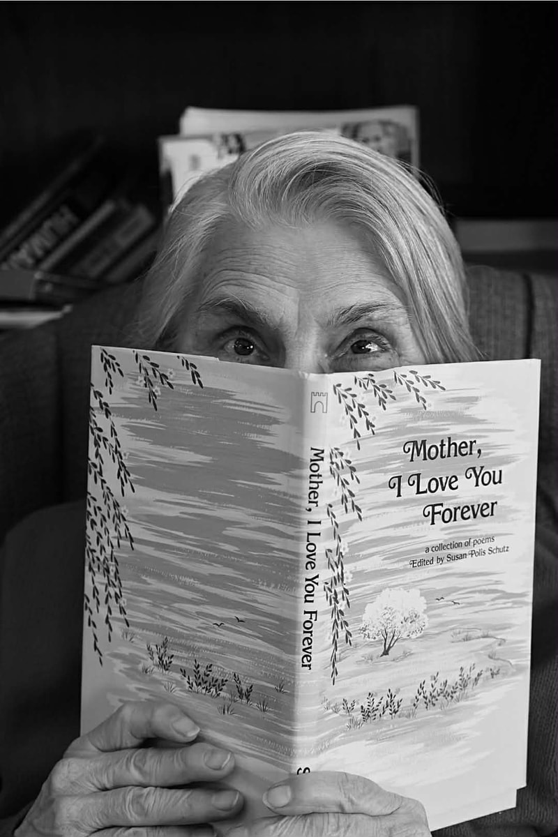 An older lady with gray hair and her eyes peeking over the top of an open book titled, Mother I love you forever.