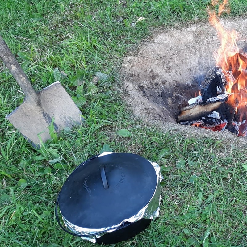 A shovel staked in the ground next to a cast iron kettle of beans ready to go into a hole in the ground. A fire burning in the hole nearby.