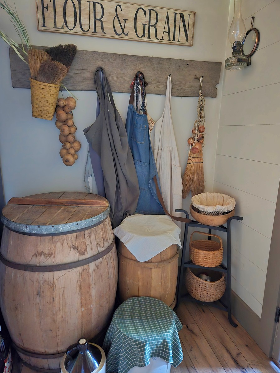 aprons hanging from an old board set with square nails. a flour & grain sign hangs above. barrels, crocks, and baskets gather on the wood floor around them