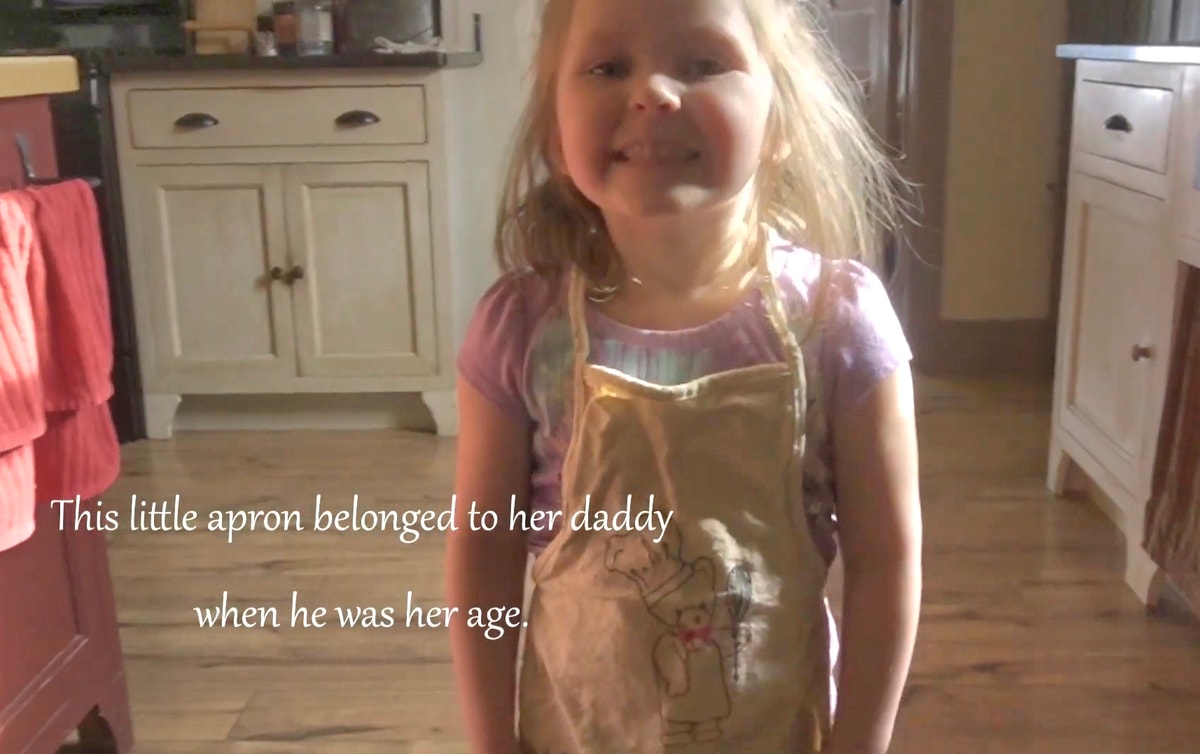 a little girl around 4 years old smiling proudly as she poses for a picture wearing the apron that once belonged to her daddy