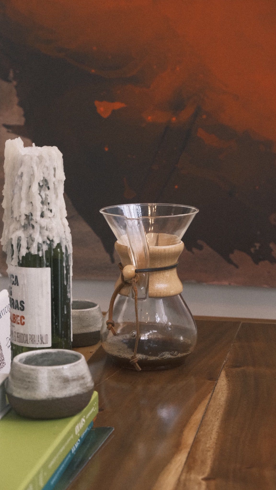 Una Chemex con café recién filtrado sobre la mesa de madera, junto a una vela en una botella y dos pocillos de cerámica