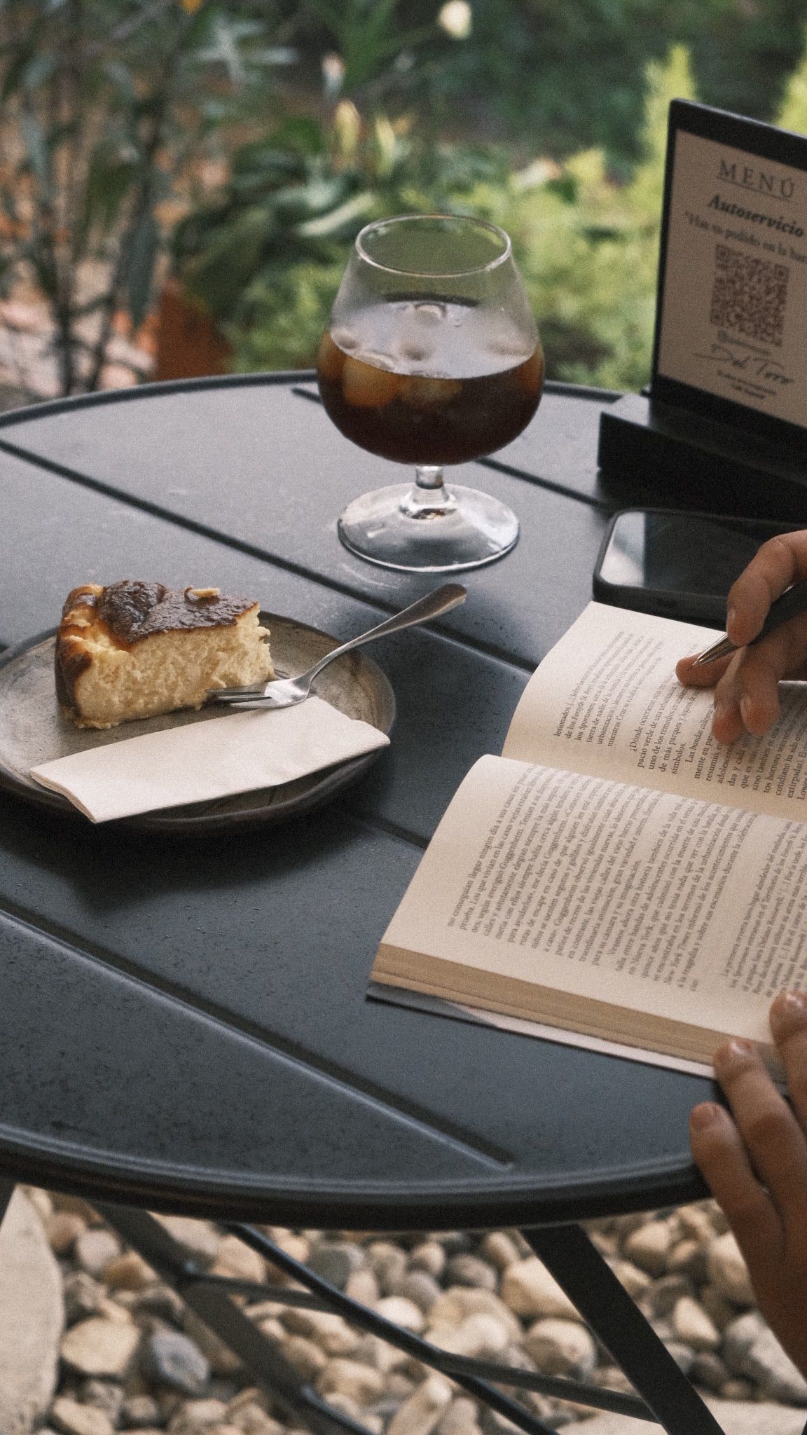Una porción de torta de queso, un café frío en copa y un libro abierto sobre una mesa del jardín