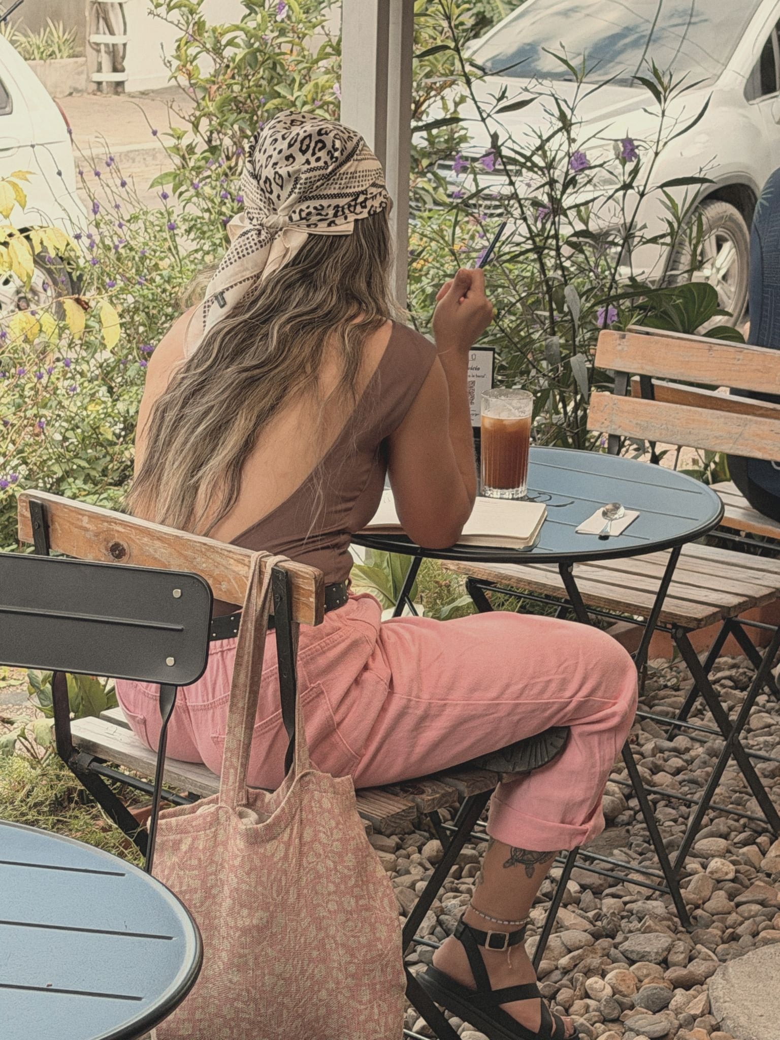 Una mujer de espaldas, con pantalón rosado, toma algo frío en una mesa azul del jardín