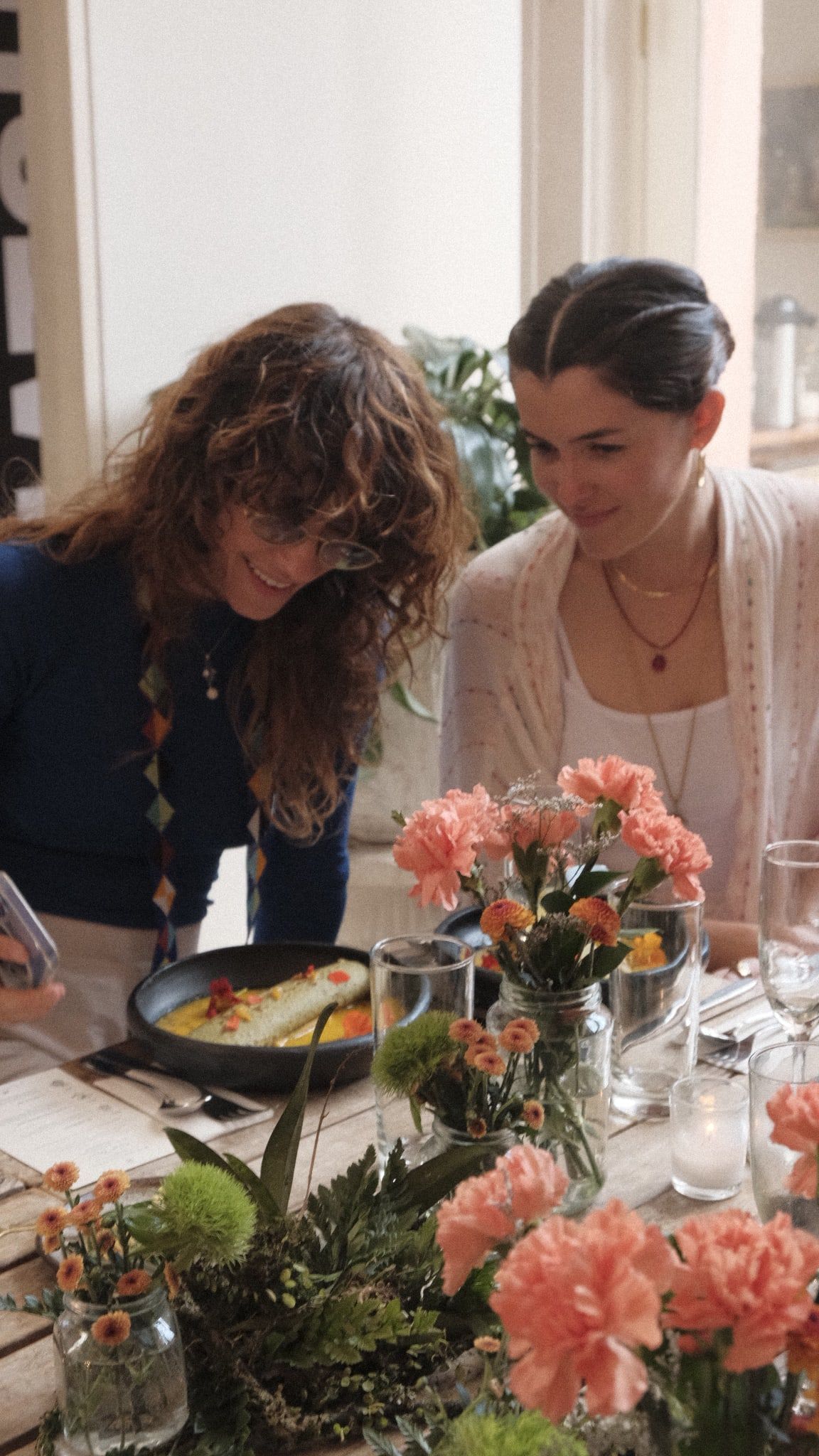 Dos invitadas miran de cerca un plato servido con flores comestibles, entre claveles y velas