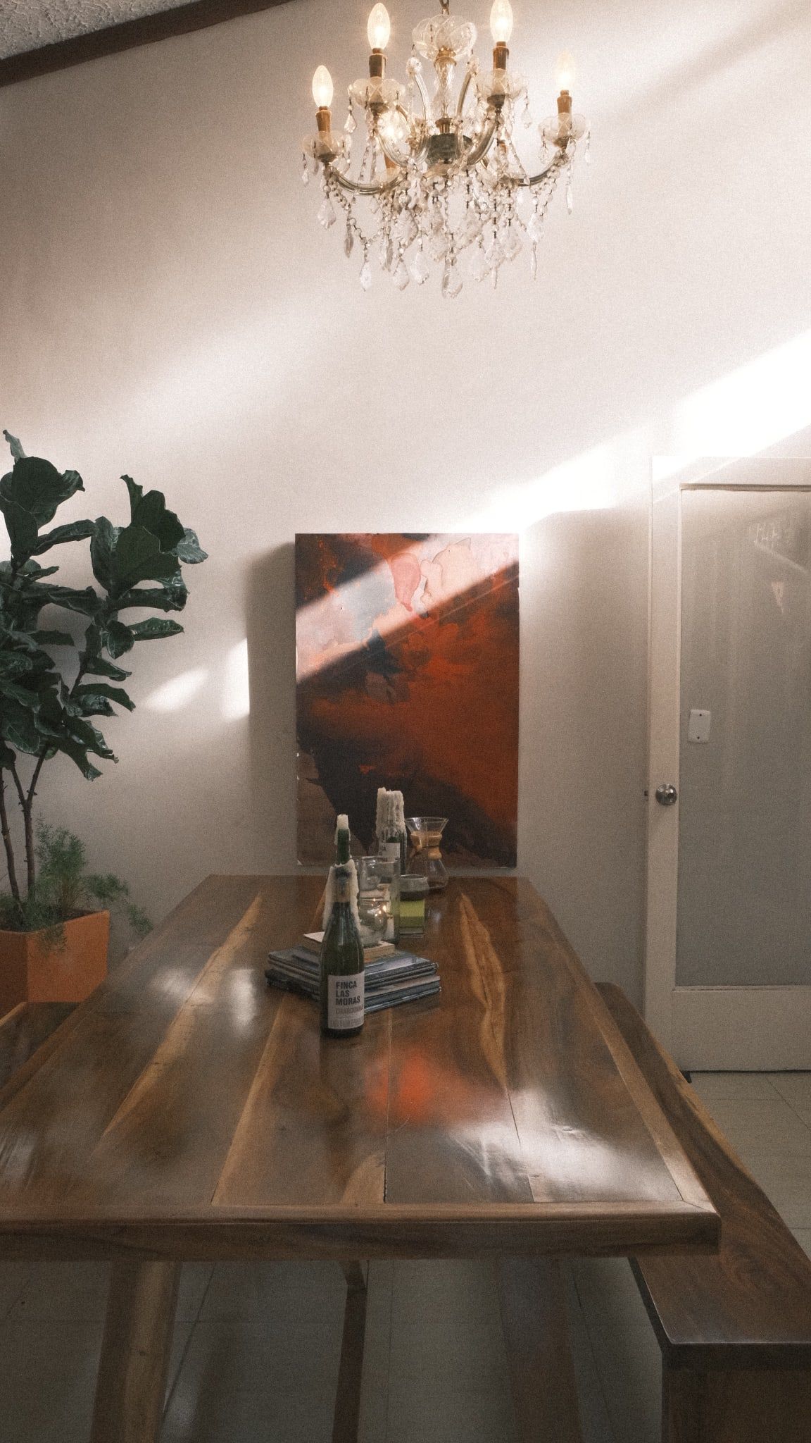 La mesa larga de madera bajo la lámpara de cristal, con un cuadro rojo apoyado en la pared y un rayo de sol