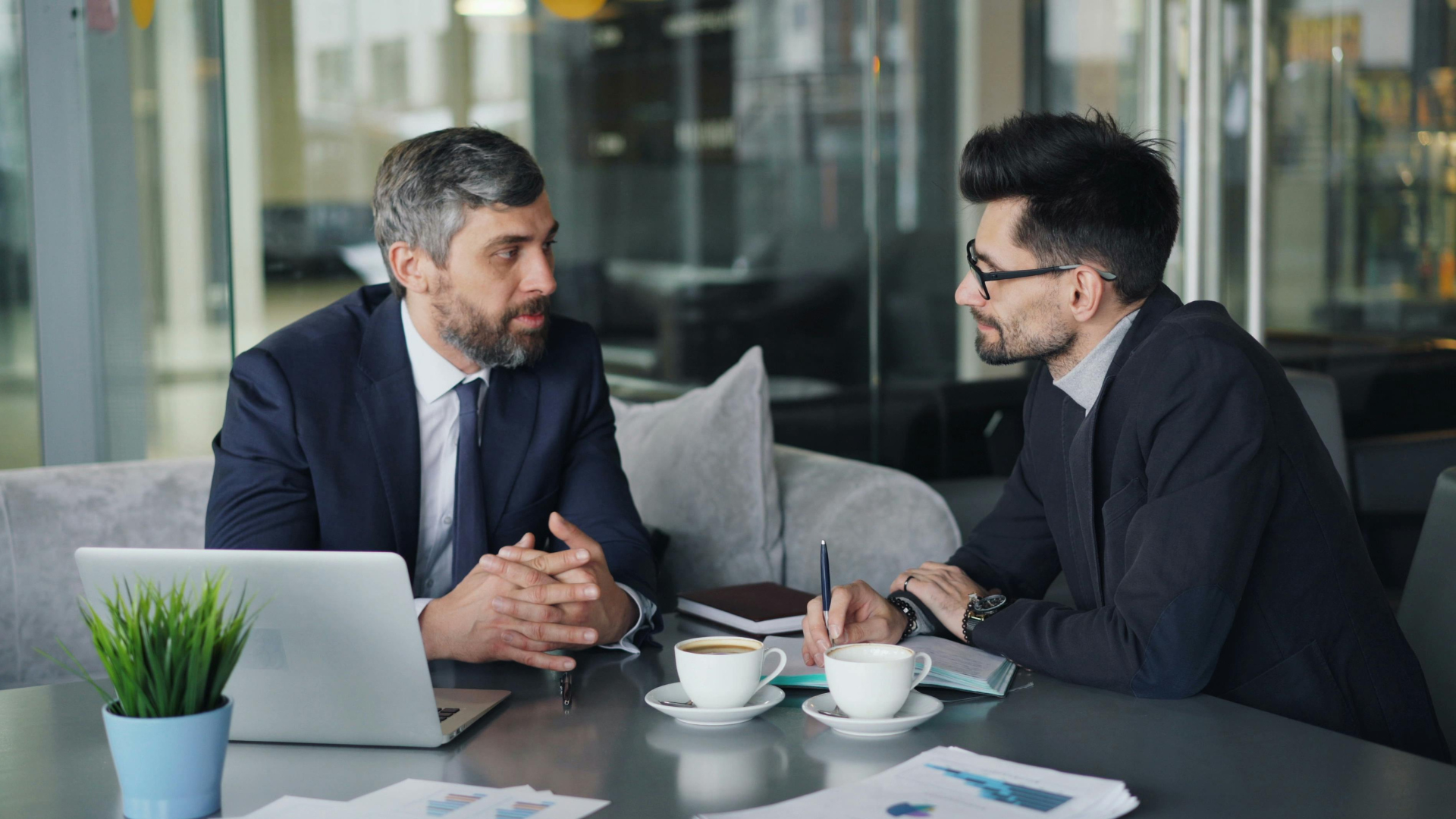 two men in suits sitting at a table with coffee cups and laptops