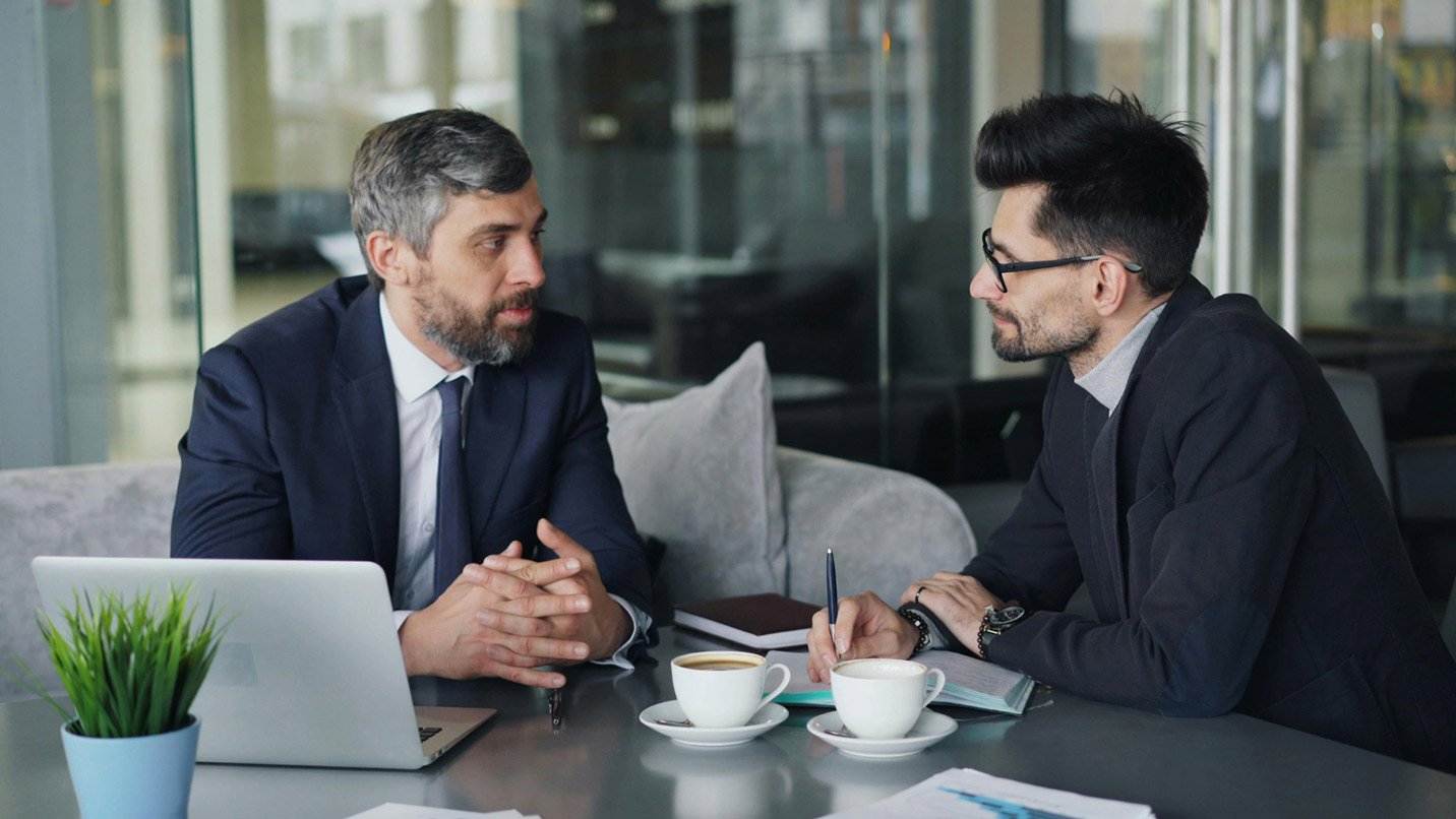 two men sitting at a table with coffee cups and a laptop