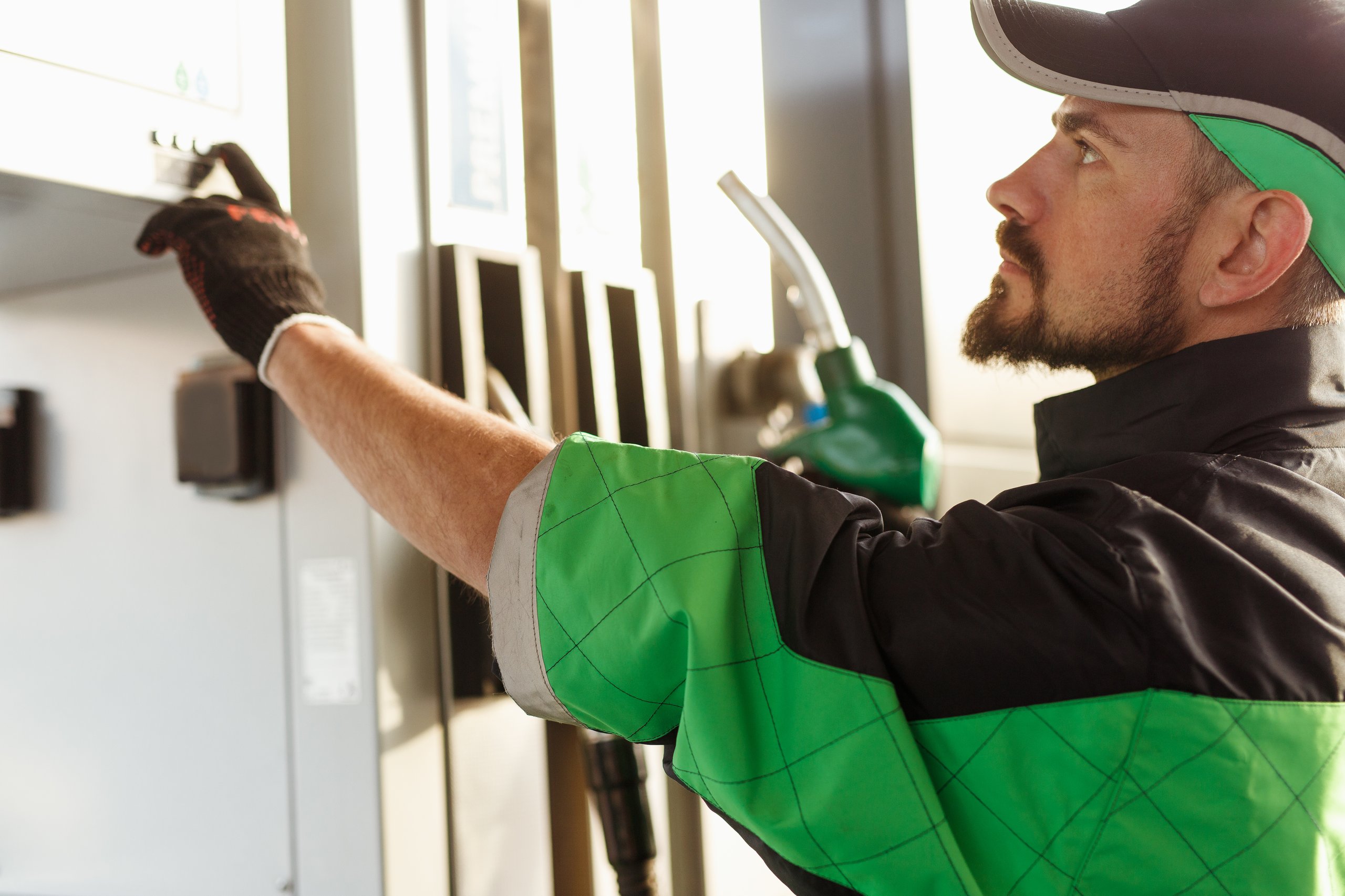 A person at a gas station reaches for the control panel of a fuel pump; a green fuel nozzle is visible in the background.