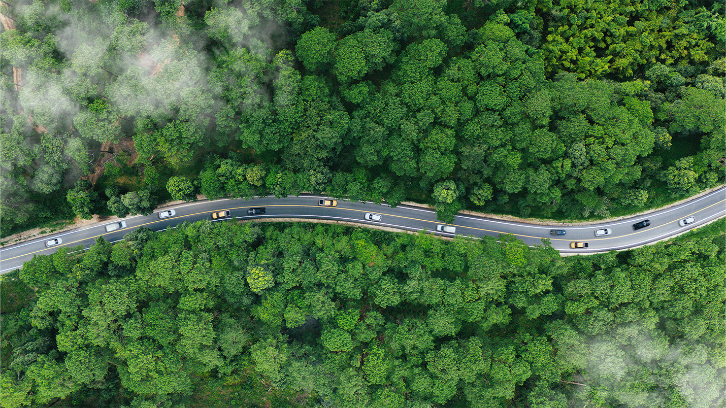 A birds eye view of a motorway surrounded by forest, with clouds above.