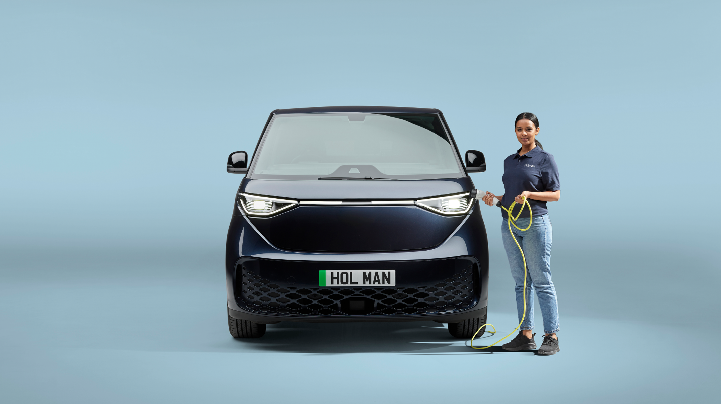 A woman holds an electric vehicle charger, stood next to an electric vehicle with a Holman number plate.