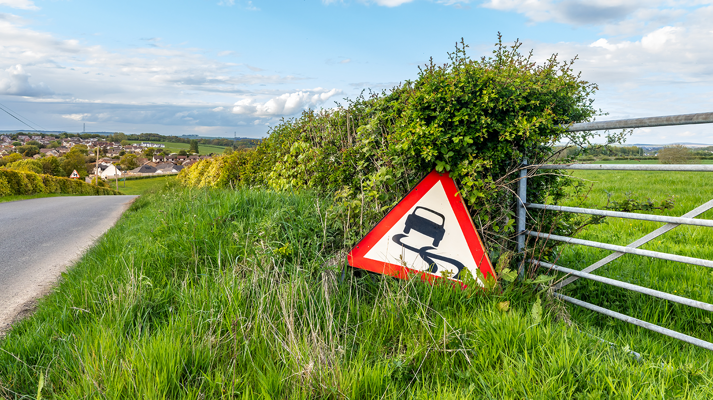 A slippery road sign placed next to a metal farm gate, next to a country lane.