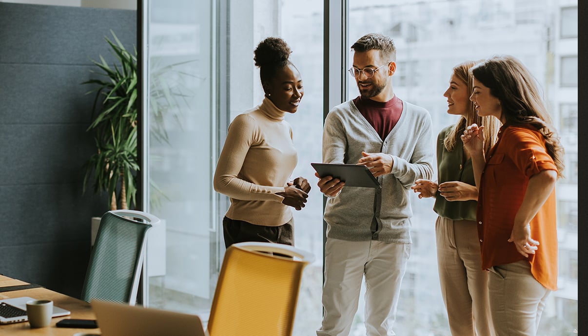 Four colleagues in a modern conference room huddled around a tablet.