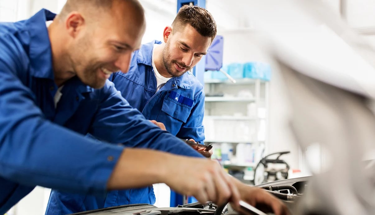 Two mechanics in blue jumpsuits working under the hood of a car.