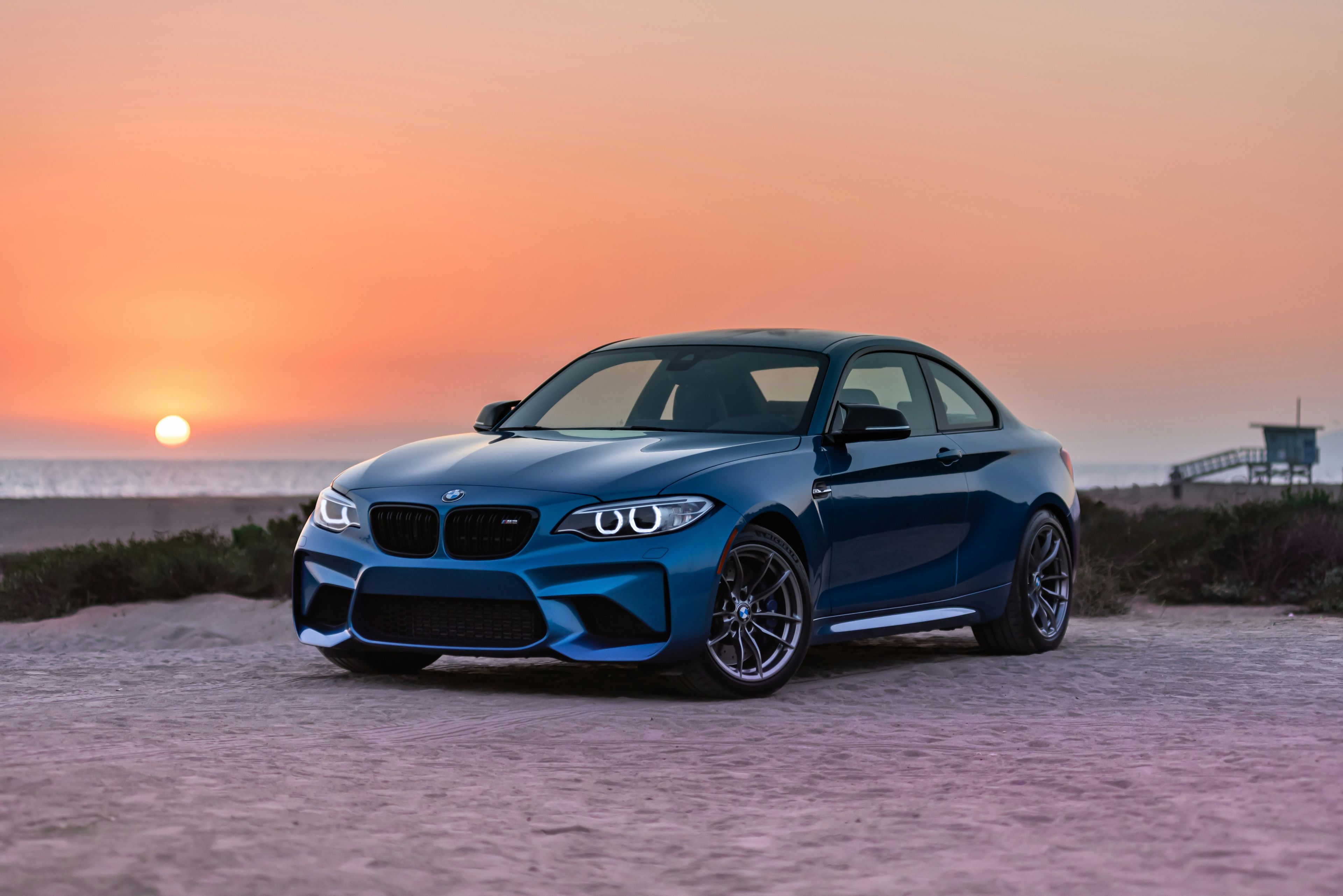 Blue BMW coupe parked on sand near a beach at sunset
