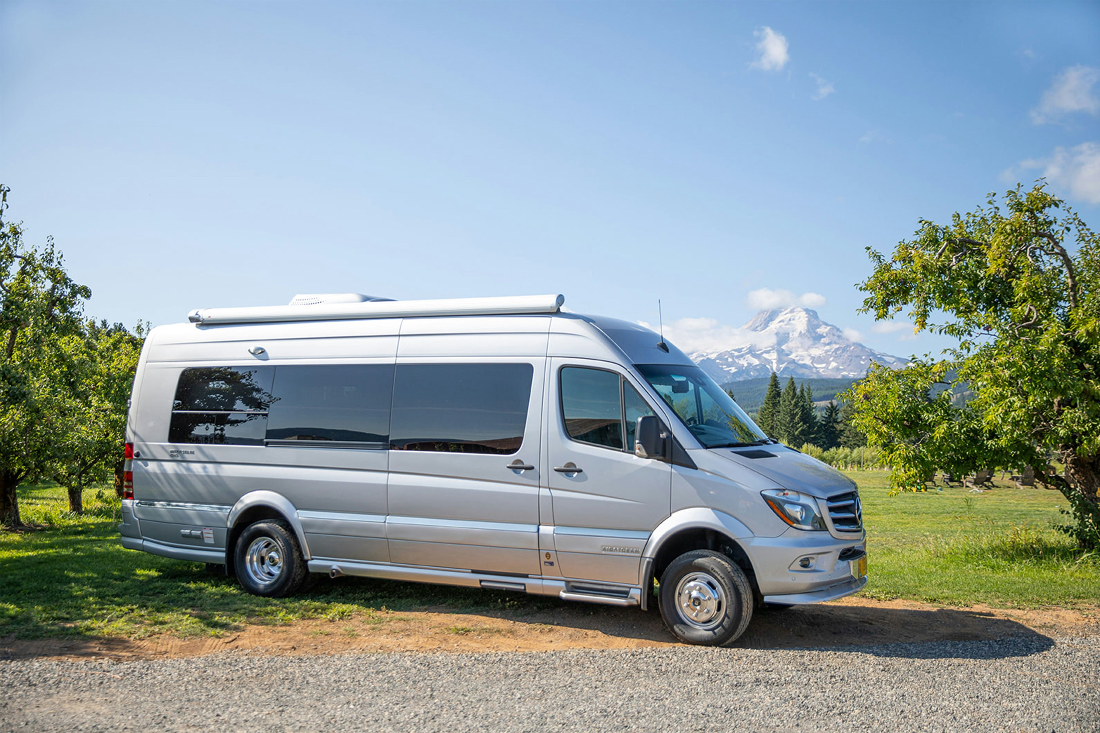 Silver Mercedes Sprinter camper parked in an orchard below a mountain