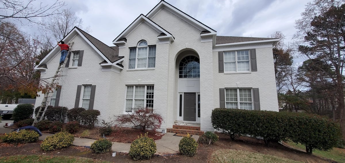 Professional painters performing final touch-ups on a white painted brick home on Debrock Court in the Preston Village neighborhood of Cary, NC, providing a technical case study for luxury exterior painting costs.