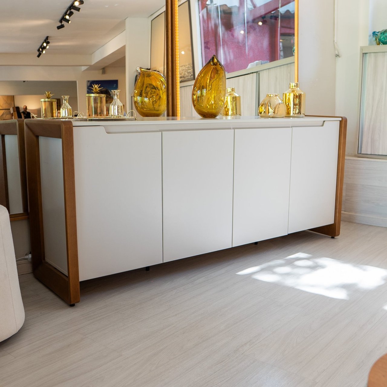 Buffet Bristol com portas brancas e moldura em madeira natural, sobre piso claro, decorado com vasos dourados