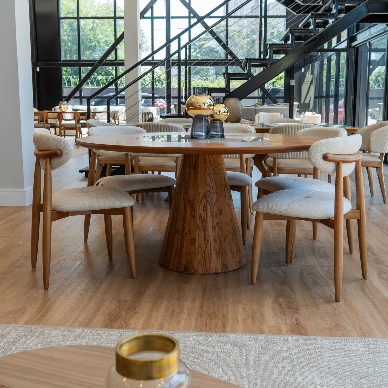 Mesa de jantar redonda Allure com tampo de madeira e base cônica maciça, cercada por cadeiras estofadas claras em showroom