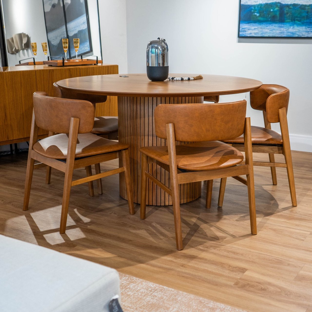 Mesa de jantar redonda Select com tampo em lâmina de madeira e base cilíndrica ripada, cercada por cadeiras de couro caramelo