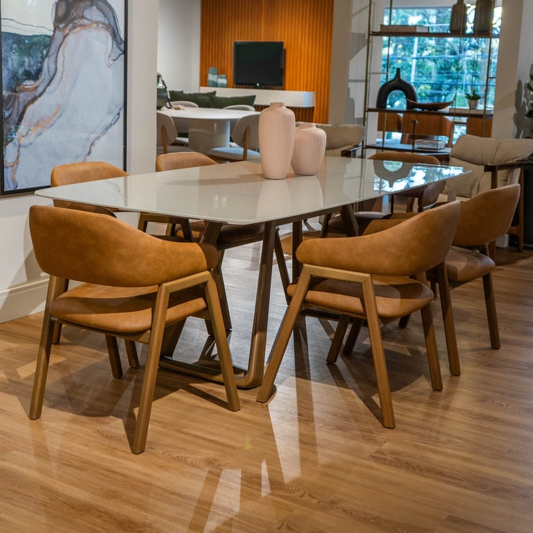 Mesa de jantar Zoe com tampo off-white e pés triangulares em madeira, rodeada por cadeiras estofadas em caramelo, em showroom