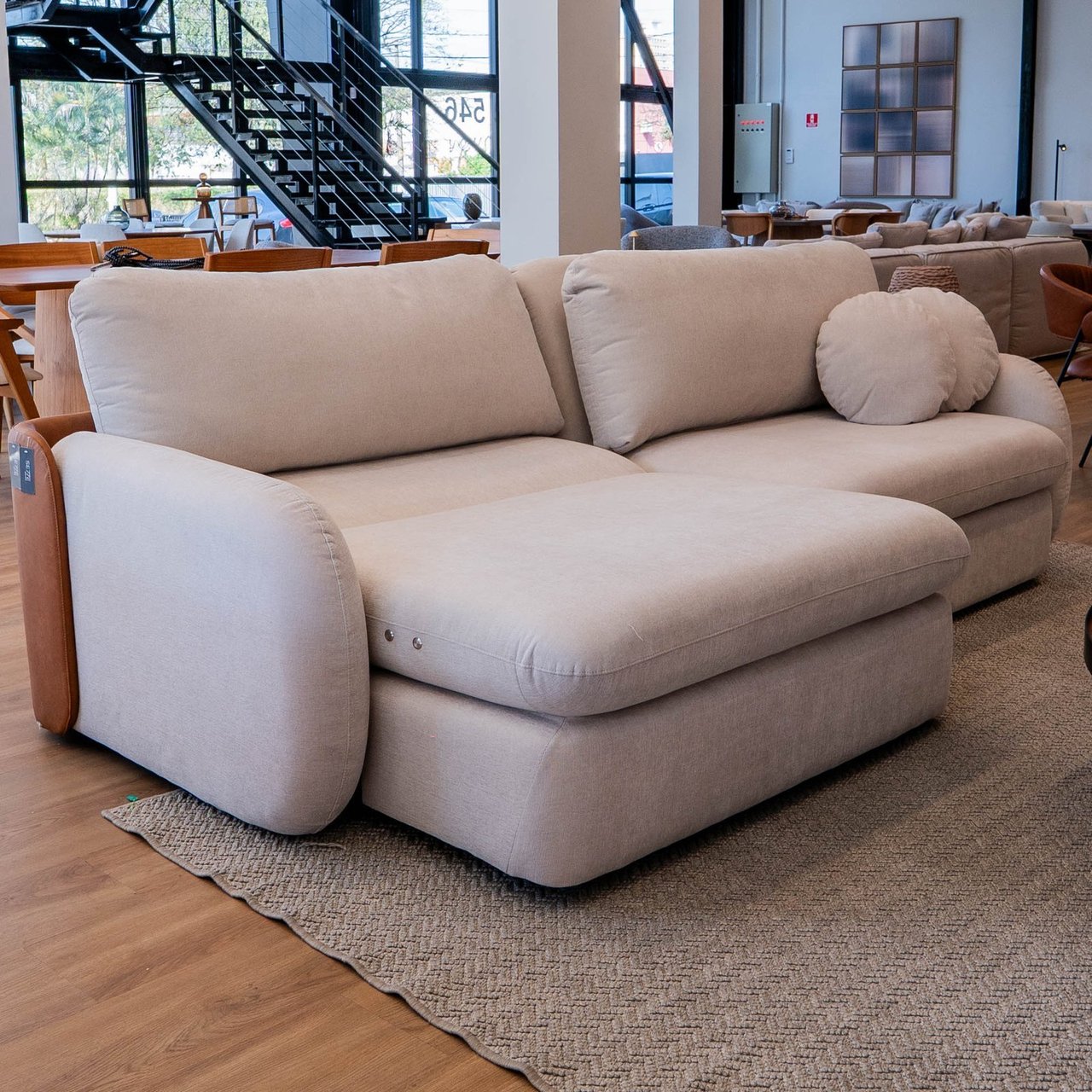 Sofá Naturalle de linhas arredondadas em tecido areia, com assento estendido e almofada-rolo lateral, em showroom
