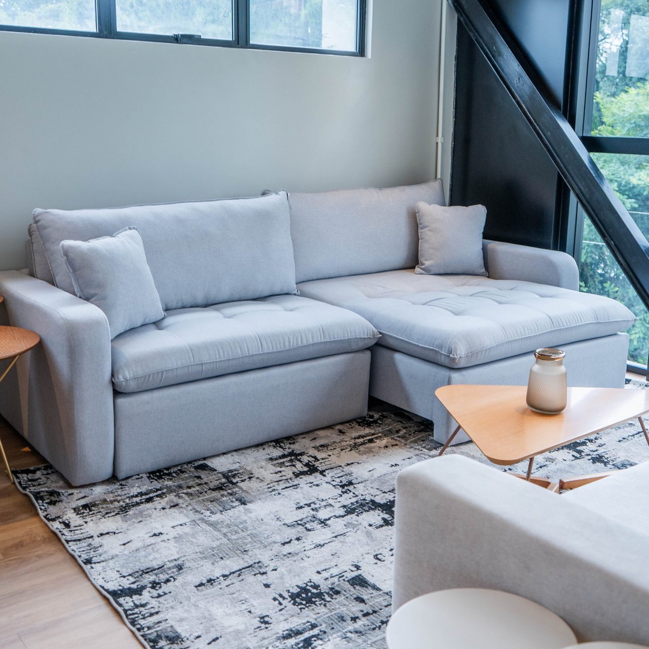 Sofá Portland em tecido cinza-claro com chaise, almofadas soltas de encosto e estrutura elevada do chão, junto a janela ampla