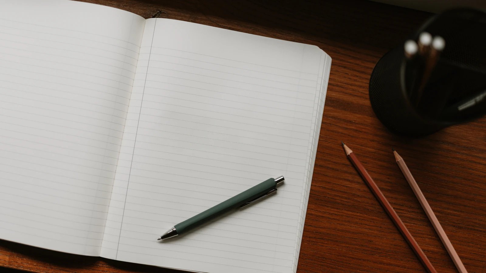An open lined notebook, a green pen and two pencils on a wooden desk.