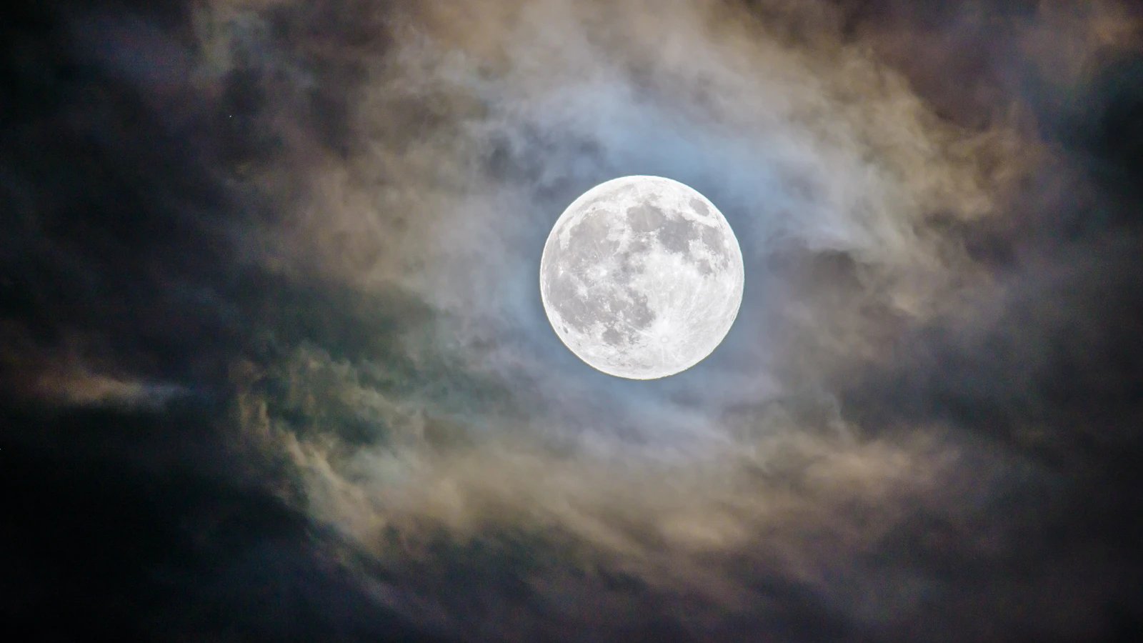 A bright full moon surrounded by softly colored clouds in a dark sky.