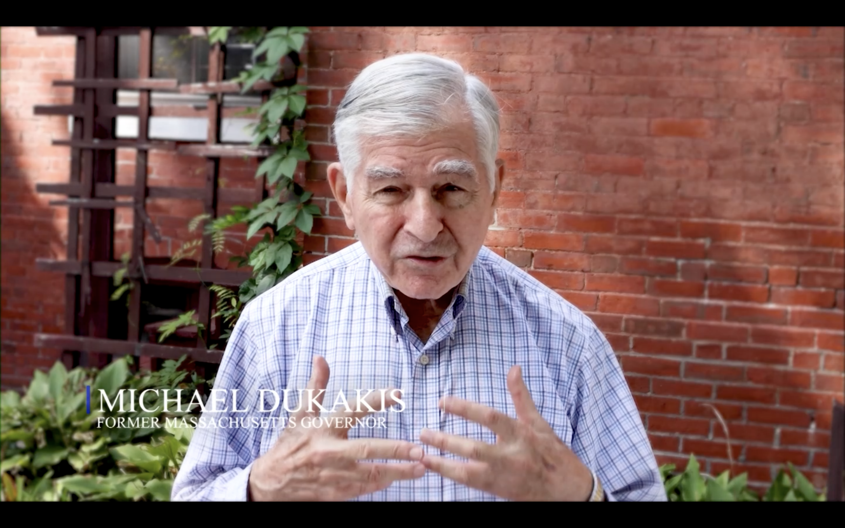 Michael Dukakis in a screenshot from one of the videos, standing in front of a red brick wall.