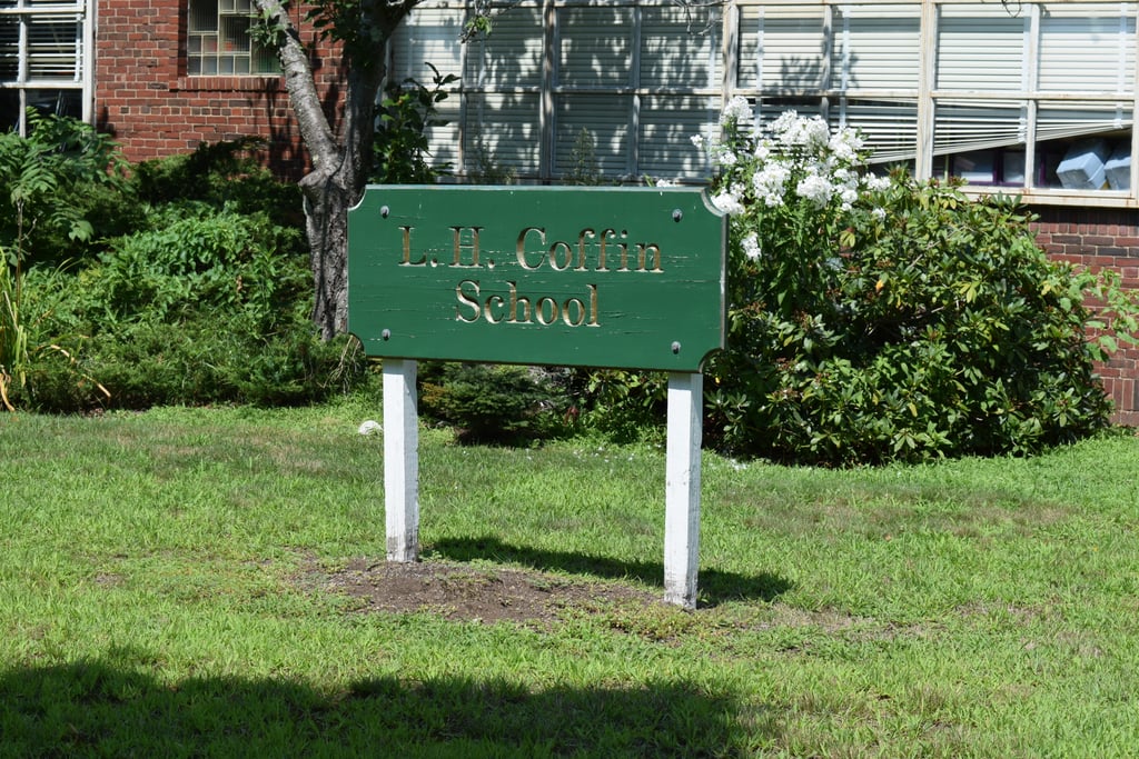 Image of former L.H. Coffin School signage. The sign sits in the foreground of the photo with foliage and the brick Coffin School building in the background. The sign is simple, of wood, in a cracked green paint with gold text inlays and held up by white posts.