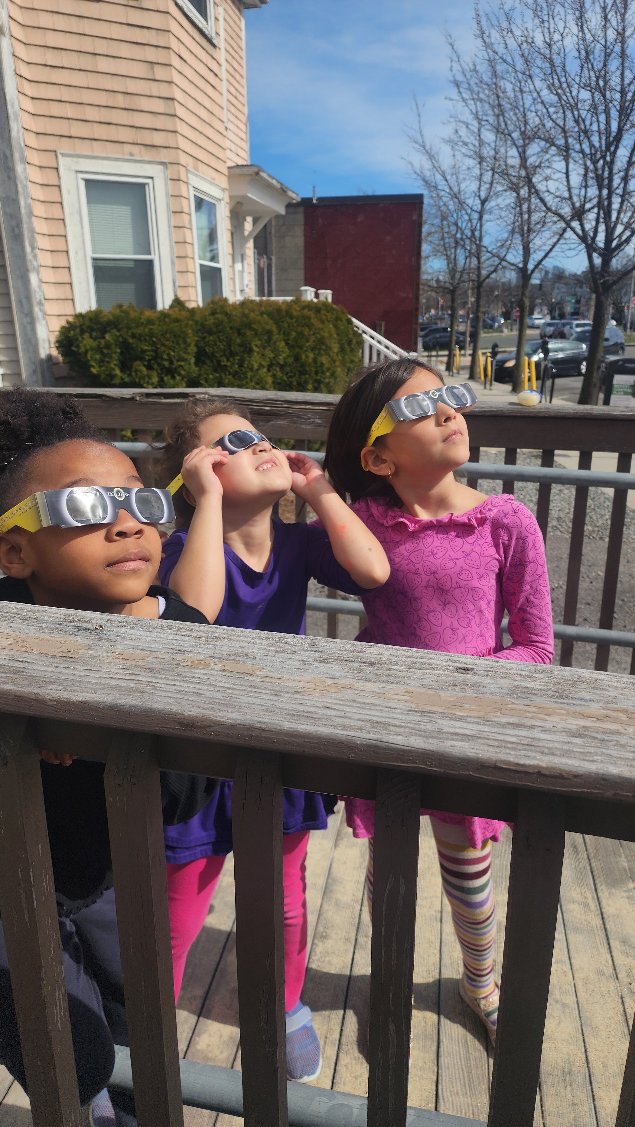 Three girls look up at the solar eclipse while wearing glasses.