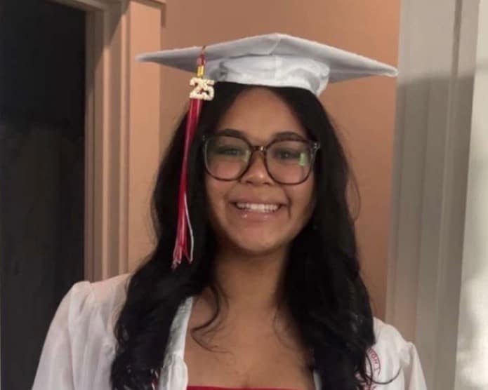Deja, a young adult with brown skin, long dark hair, and glasses wearing a white cap and gown with a red tassle
