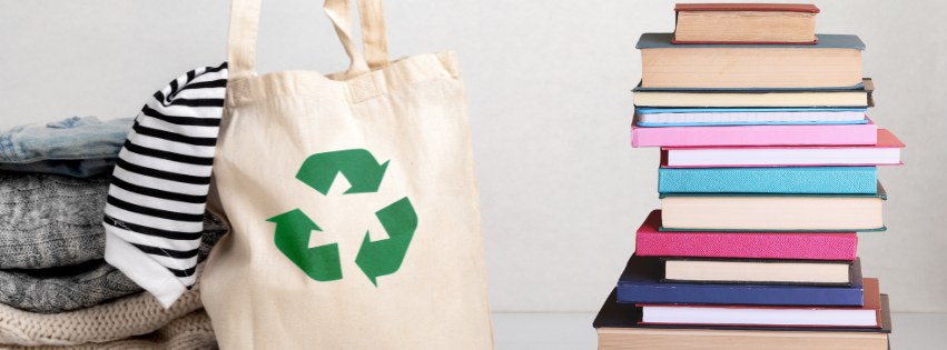 canvas colored tote bag with a green recycle logo on it filled with clothing, stack of clothing next to the bag and a stack of books