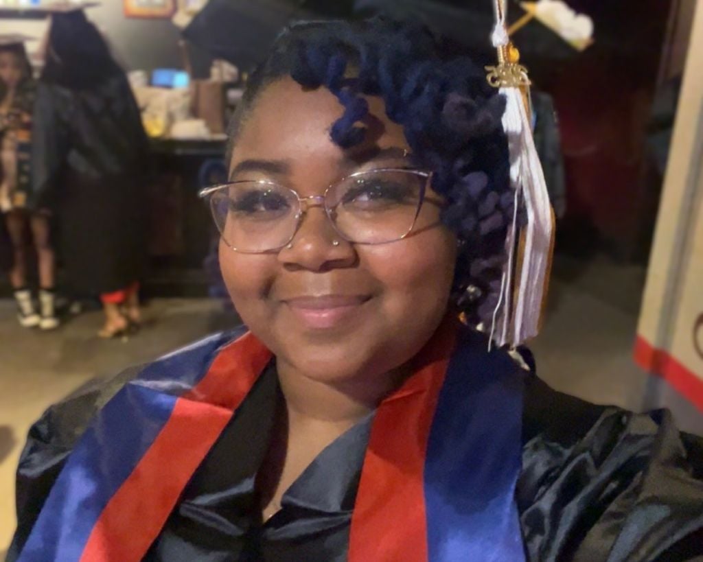 Natavia (Tavi) Boquet-Sheers, a young woman with brown skin and curly, dark brown hair wearing glasses, a black cap and gown, and a blue and red stole. 