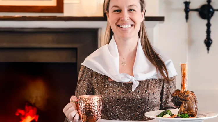 A woman wearing historical garb holds a plate and a mug.