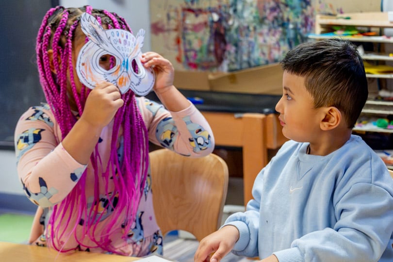Two preschoolers sharing a desk. One is showing off a paper owl mask made during a craft project