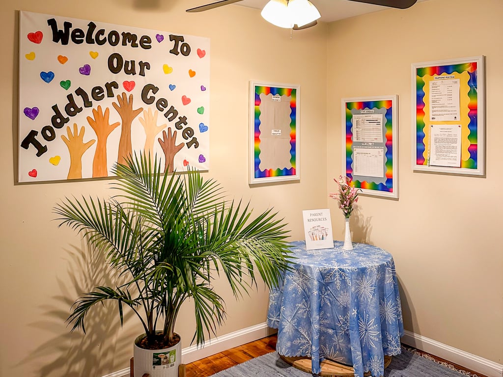 A white sign that reads "Welcome to Our Toddler Center" with multicolor hearts & hands hangs on  a warm wall above a potted palm frond and a small round table covered by a blue cloth. 