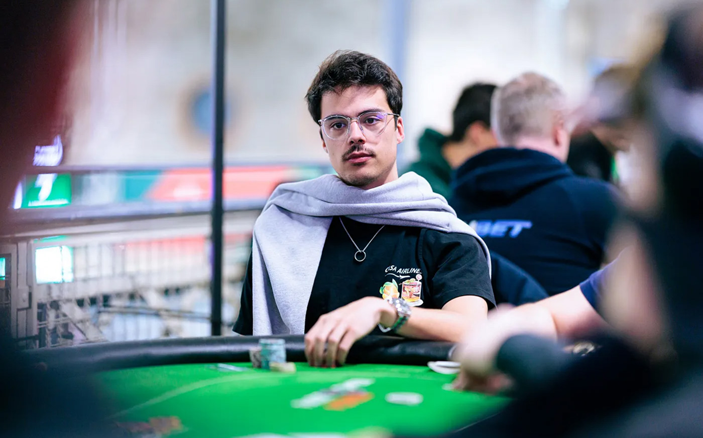 Gilles Simon seated at the table in a black T-shirt and glasses, focused during a live tournament with other players in the background.