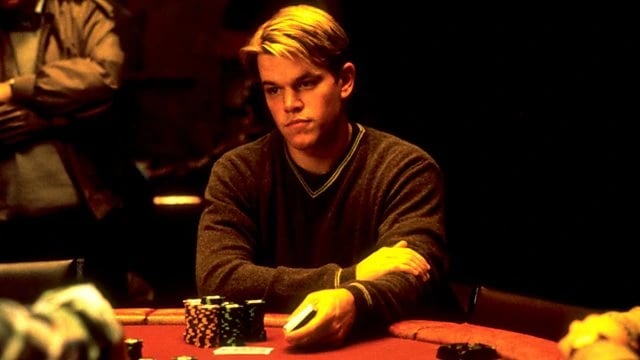 Matt Damon sits at a dimly lit poker table holding cards with stacks of chips in front of him during a tense scene from the film Rounders.