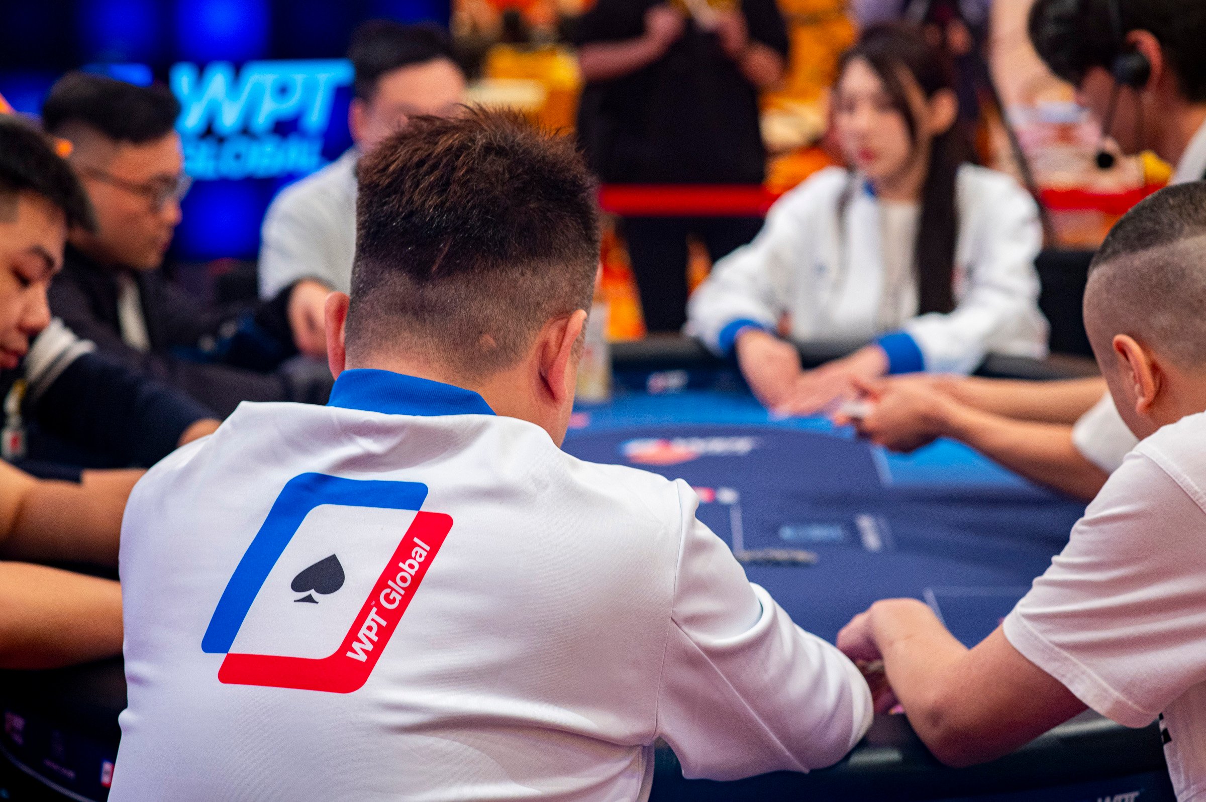 Players compete at a WPT Global poker table, wearing branded white jackets during a live tournament.