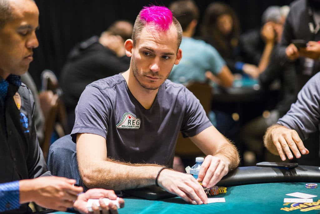 Justin Bonomo with bright pink hair concentrates during a live poker tournament, holding cards at the table.