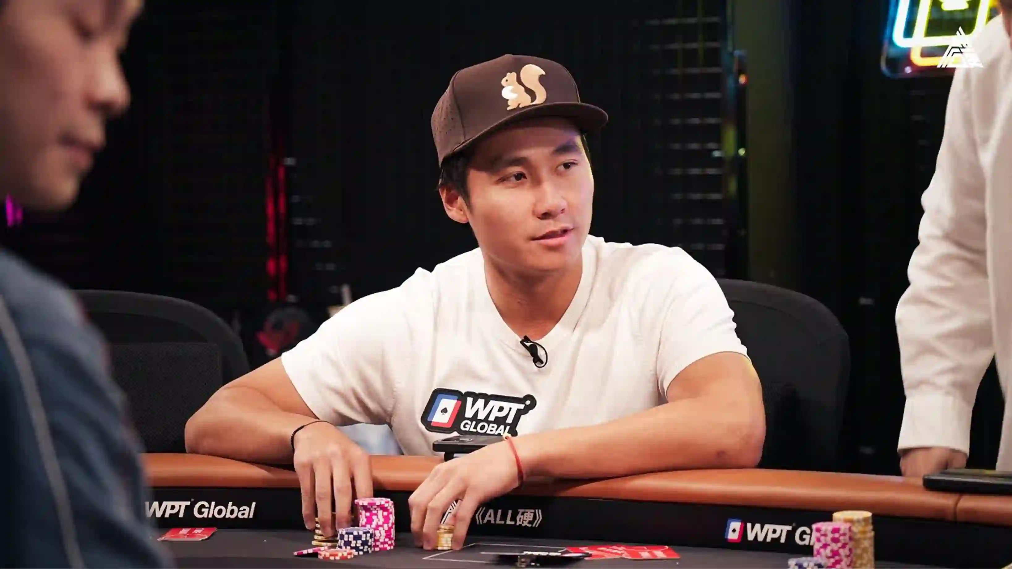 Ethan ‘Rampage’ Yau plays at a WPT Global–branded table, wearing a white shirt and brown squirrel cap with chips in front of him.
