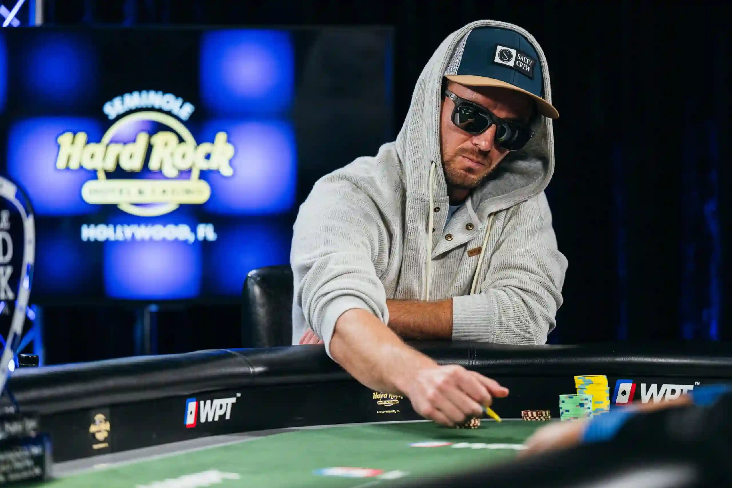 Dylan Smith in a grey hoodie and cap making a bet at a WPT® table with the Seminole Hard Rock logo on the screen behind him.